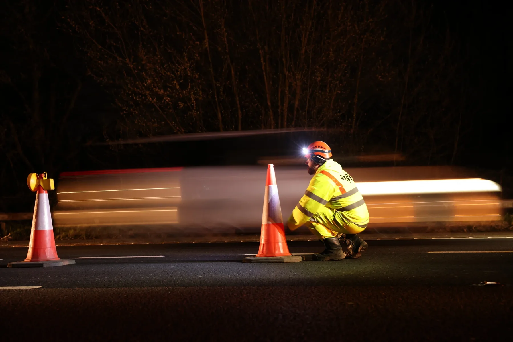 Night-time traffic management operative kneeling beside a cone with light trails from passing traffic