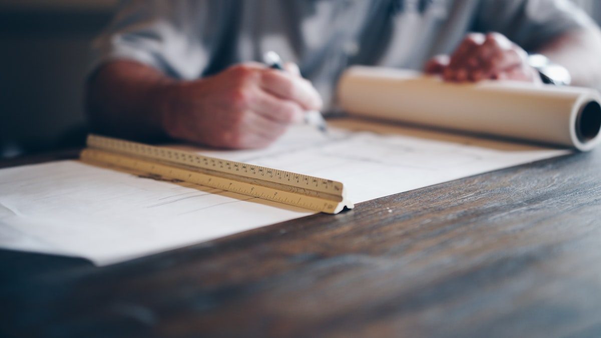 Designer sketching layouts at a wooden desk with a scale ruler and rolled artwork